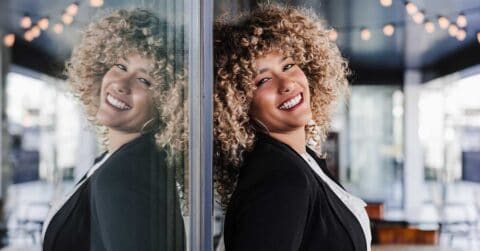 A person with curly hair exudes authentic confidence while smiling and leaning against a glass wall, their reflection visible. The indoor background is adorned with string lights.