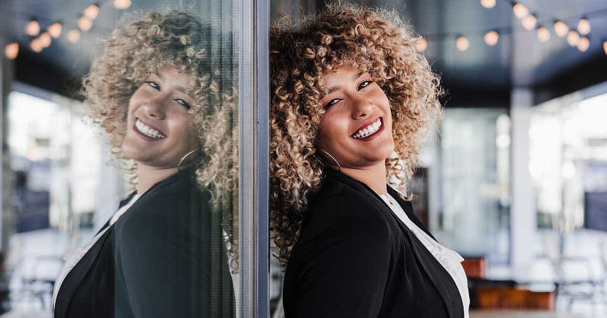 A person with curly hair exudes authentic confidence while smiling and leaning against a glass wall, their reflection visible. The indoor background is adorned with string lights.