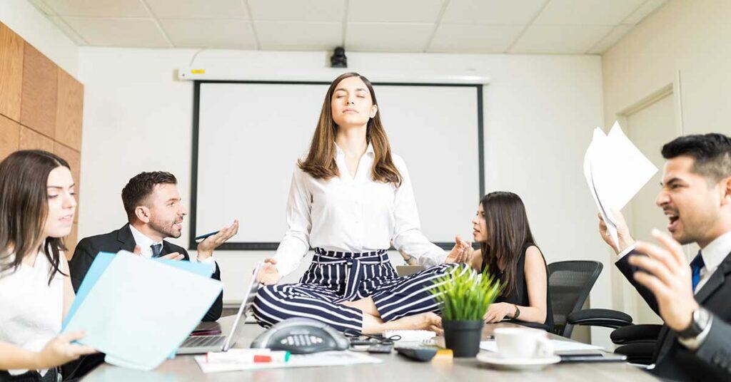 A woman sits cross-legged meditating on a conference table, embodying work-life balance as four coworkers around her appear stressed and engaged in a heated discussion.