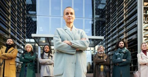 A group of seven people stand confidently with arms crossed in front of a modern glass building. The person in front wears a light coat and appears to be in a leadership position for the group.