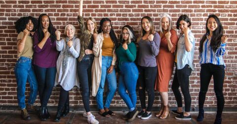 Ten women of diverse ages and backgrounds stand in a row against a brick wall, smiling with confidence and raising their fists in a gesture of empowerment.