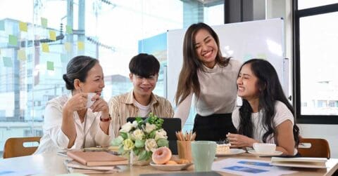 Four people sit and stand around a table in an office, smiling and talking, clearly having it as they enjoy papers, flowers, and snacks in front of them.