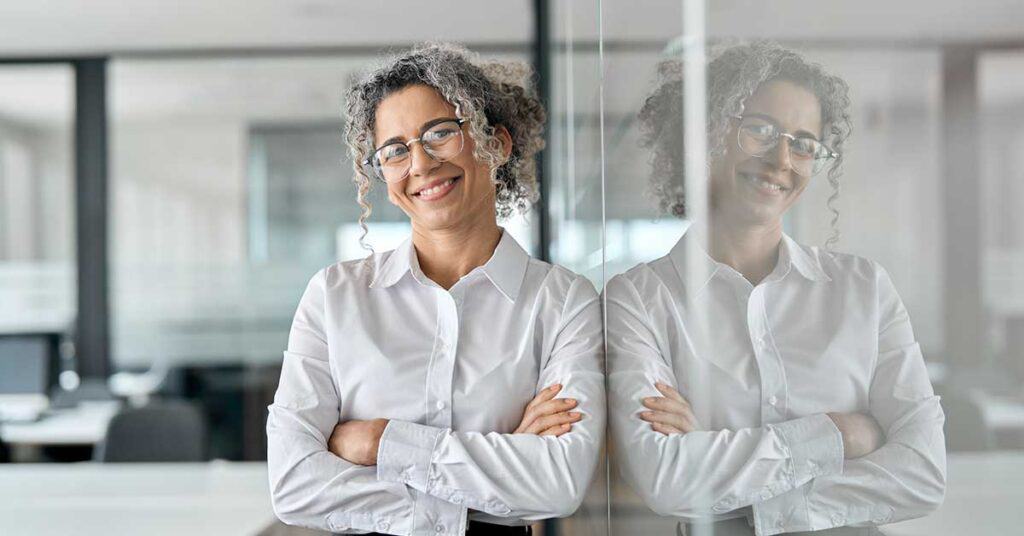 A woman with curly gray hair and glasses stands with arms crossed, smiling and confidently leaning against a glass wall in an office setting, embodying professionalism and clear boundaries.