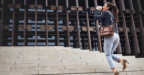 A woman in business attire walks up stone steps, holding a cup and looking ahead, symbolizing progress against the backdrop of a modern building with a grid-like facade.