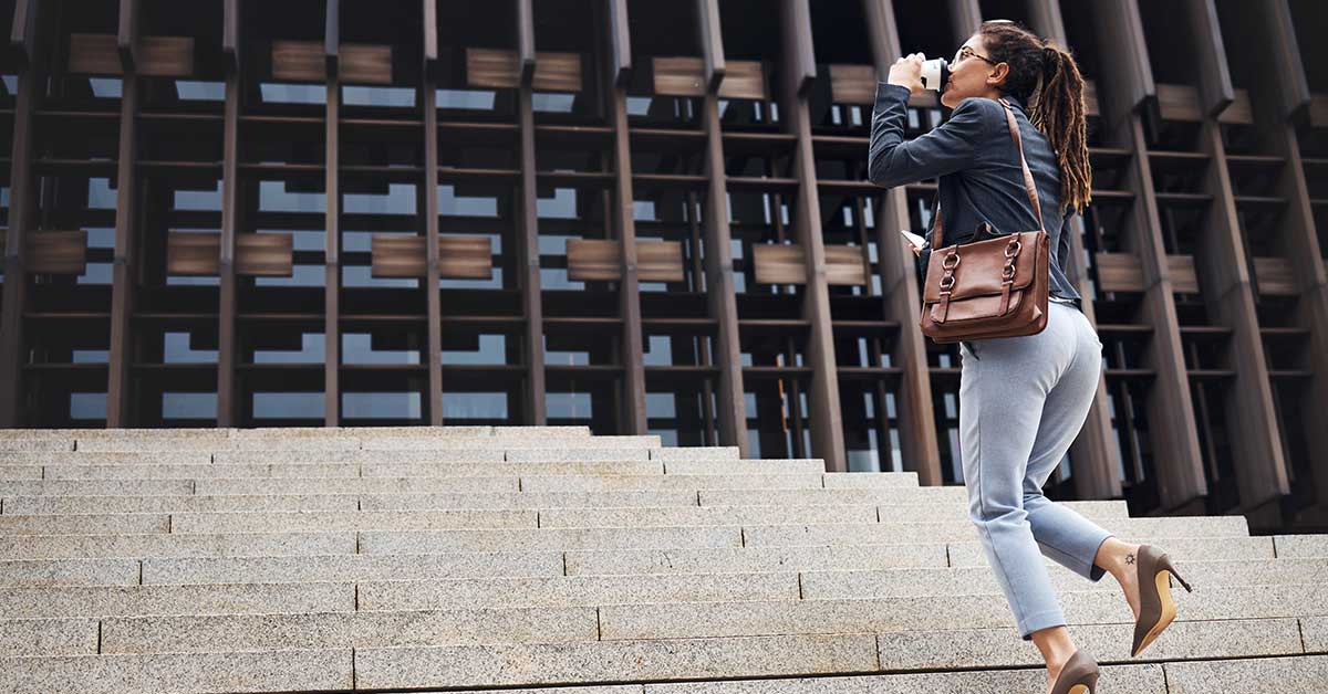 A woman in business attire walks up stone steps, holding a cup and looking ahead, symbolizing progress against the backdrop of a modern building with a grid-like facade.