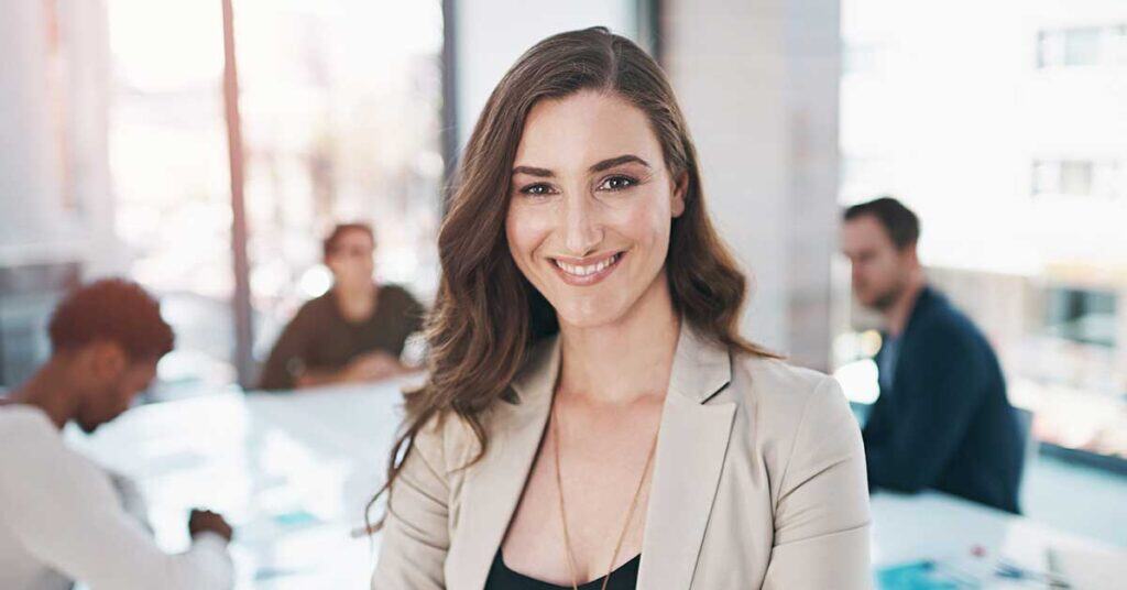 A woman in business attire, exemplifying authentic leadership, smiles at the camera in a modern office, while three colleagues work in the background.