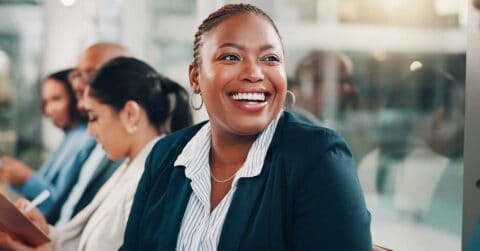 A woman in business attire smiles imperfectly while seated at a conference table with colleagues in a professional setting.
