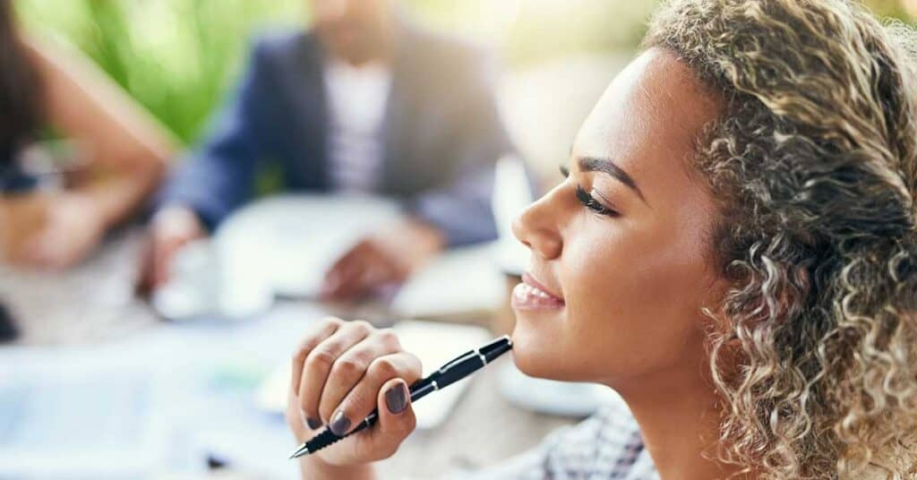 A person with curly hair holds a pen to their chin and smiles thoughtfully, embodying the spirit of being intentional, with blurred people and papers in the background.