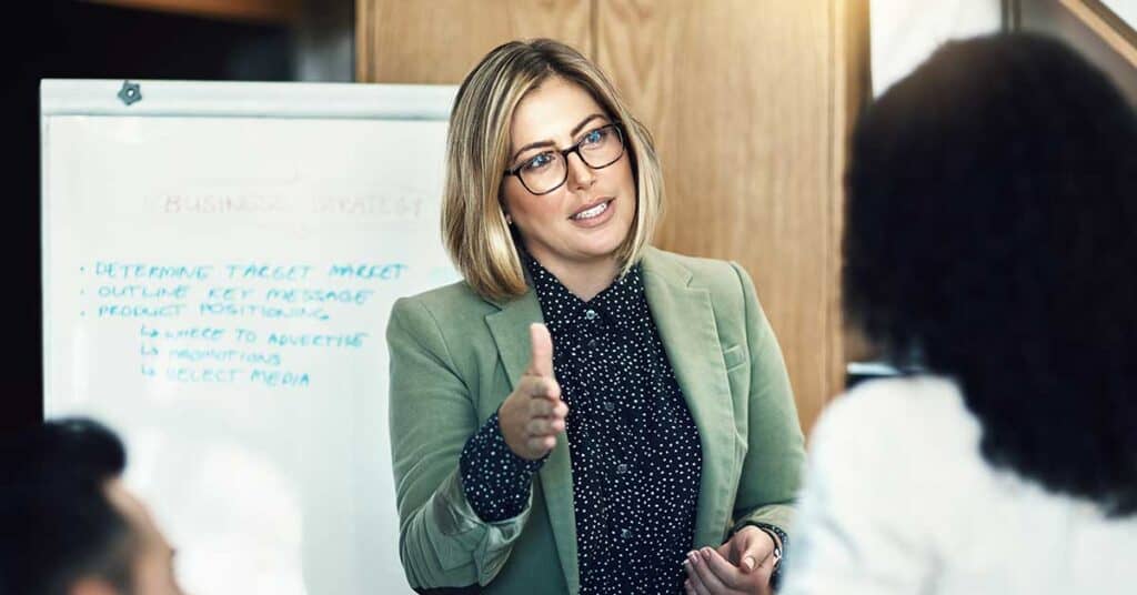A woman in glasses and a green blazer leads a coaching session, gesturing as she speaks in front of a whiteboard with business notes to two colleagues.