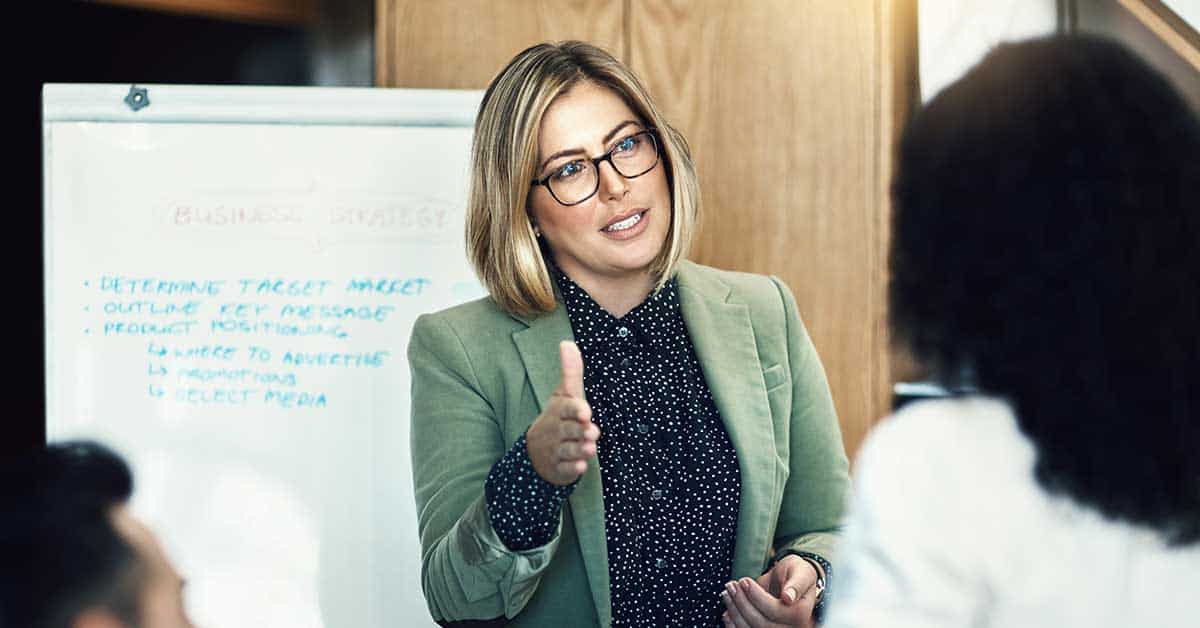 A woman in glasses and a green blazer leads a coaching session, gesturing as she speaks in front of a whiteboard with business notes to two colleagues.