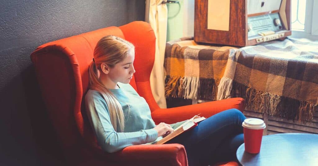 A woman sits in a red armchair reading a book, with a takeaway coffee cup on the table nearby and a vintage radio in the background, taking time to invest in herself through quiet moments.