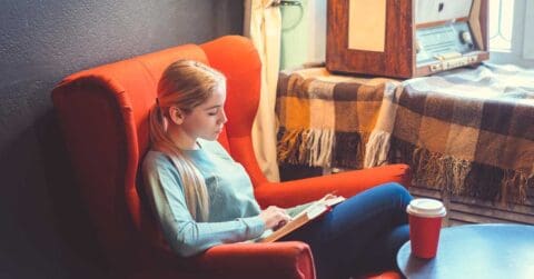 A woman sits in a red armchair reading a book, with a takeaway coffee cup on the table nearby and a vintage radio in the background, taking time to invest in herself through quiet moments.