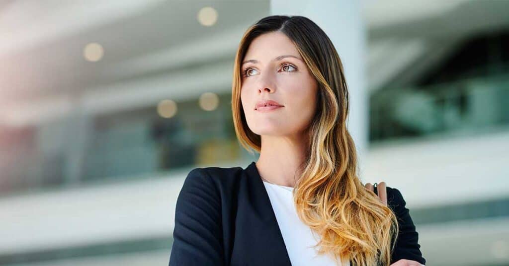A woman with long, wavy hair wearing a dark blazer and white shirt stands indoors, looking slightly to the side with a neutral expression that hints at quiet self-worth.
