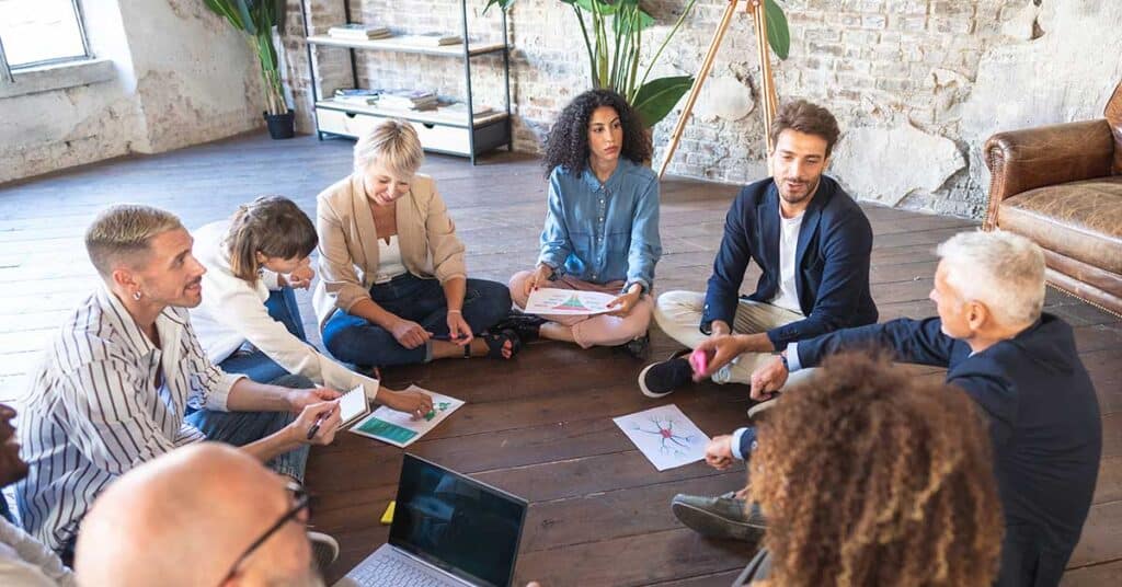 A group of people sit in a circle on a wooden floor, discussing documents and charts in a casual, village-inspired loft-style room.