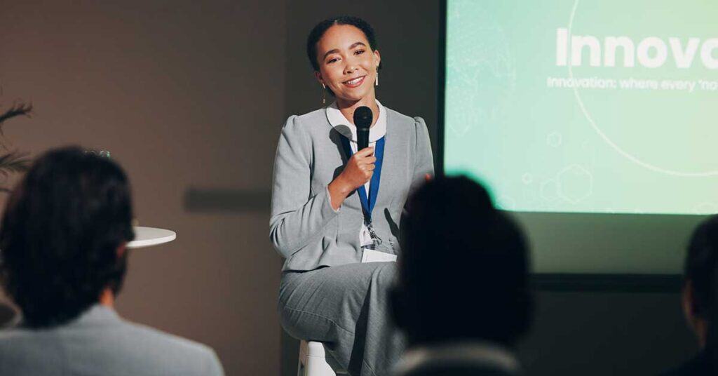 A woman in a gray suit holds a microphone and addresses the audience during a presentation on imposter syndrome, with a screen displaying the word "Innov" in the background.