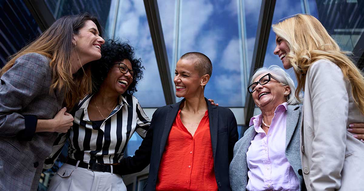Five women of diverse ages and styles stand close together outside, smiling and talking in front of a glass building with blue sky reflected in the windows, radiating a transformational sense of unity and empowerment.