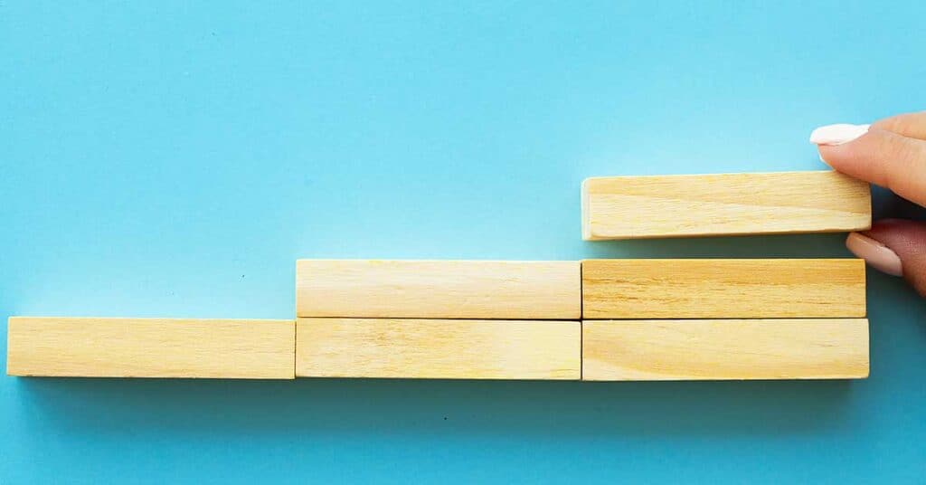 A hand places a rectangular wooden block on top of a row of blocks, symbolizing change as it forms a stepped structure against a blue background.