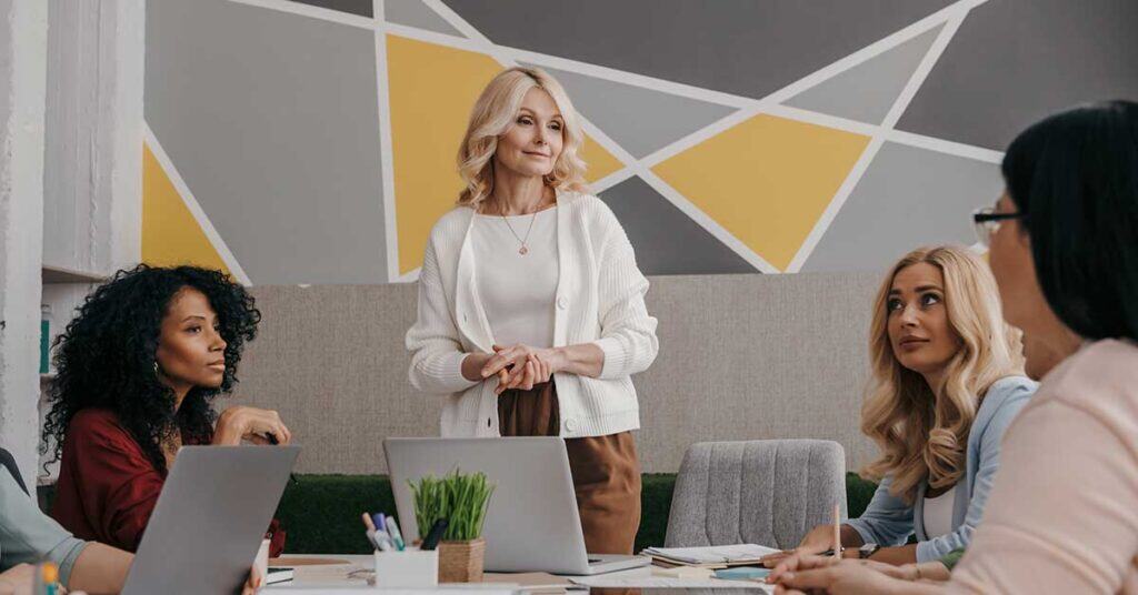 A woman stands and speaks to three seated women during a meeting in a modern office, demonstrating leadership development as they collaborate with laptops and documents on the table.