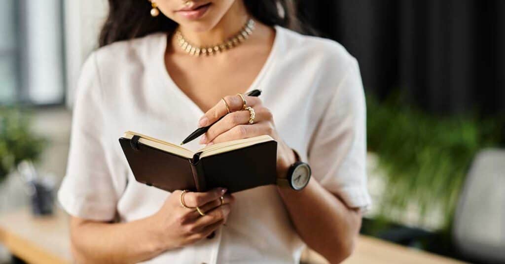 A woman wearing a white blouse and jewelry writes in a small notebook while standing indoors, capturing her thoughts on professional growth.