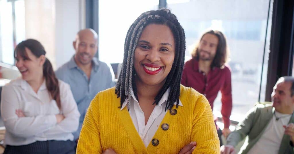 A woman in a yellow sweater stands smiling in the foreground, embodying resilience, while four people sit and talk in the background of a bright office setting.