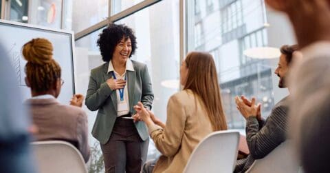 A woman stands and smiles while showcasing her skill as she presents to a group of people who are seated and applauding in a modern office setting.