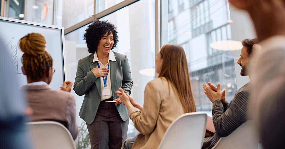 A woman stands and smiles while showcasing her skill as she presents to a group of people who are seated and applauding in a modern office setting.