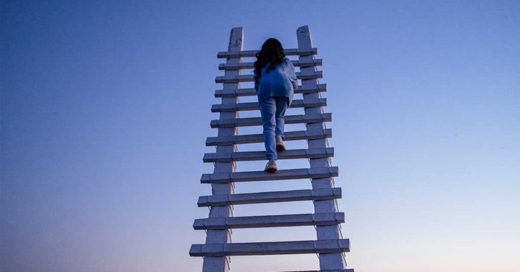 A person climbs a tall white wooden ladder with a broken rung against a clear blue sky at dusk.