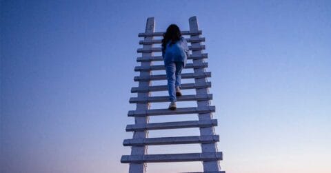 A person climbs a tall white wooden ladder with a broken rung against a clear blue sky at dusk.