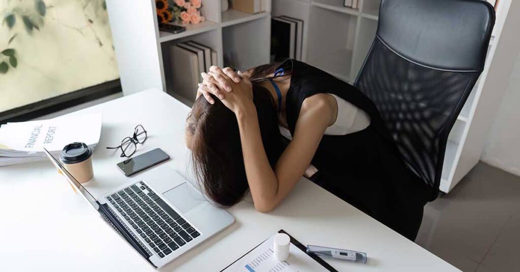 A person sits at a desk with head down and hands on their head, surrounded by a laptop, documents, phone, coffee cup, and office supplies—highlighting the pressures that can come with leadership.
