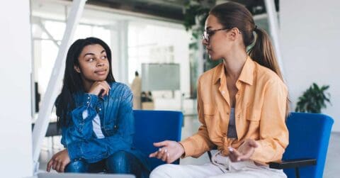 Two women sit in an office space, engaged in a conversation about leadership. One woman gestures with her hands while the other listens attentively.