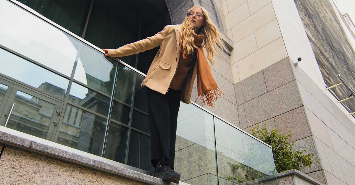 A woman with long blonde hair, wearing glasses, a beige coat, and scarf stands on a stone ledge next to a glass railing outside a modern building, subtly capturing the concept of the glass cliff.