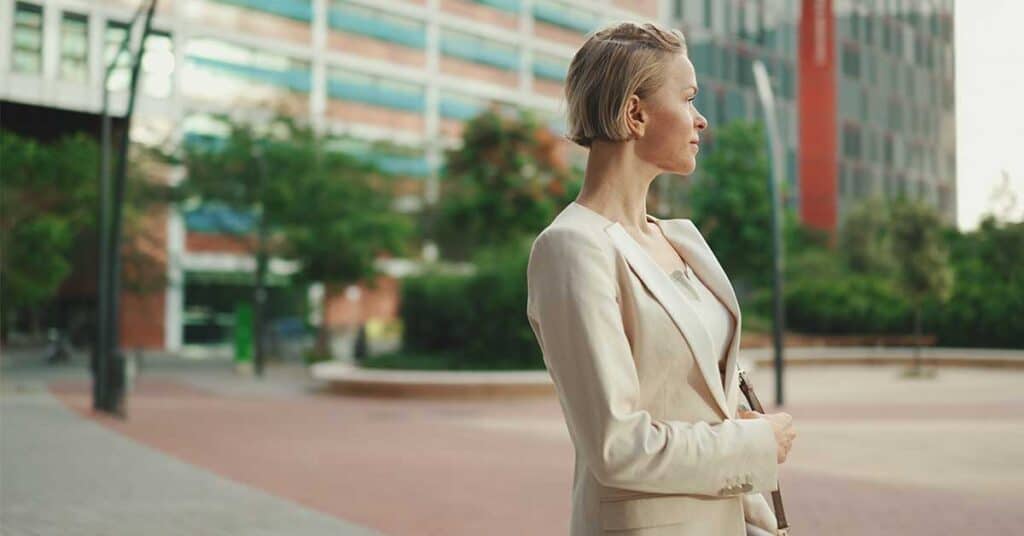 A stylish woman in a light-colored suit stands outdoors in an urban setting, looking to the side. The scene captures confident women against a backdrop of buildings and trees.