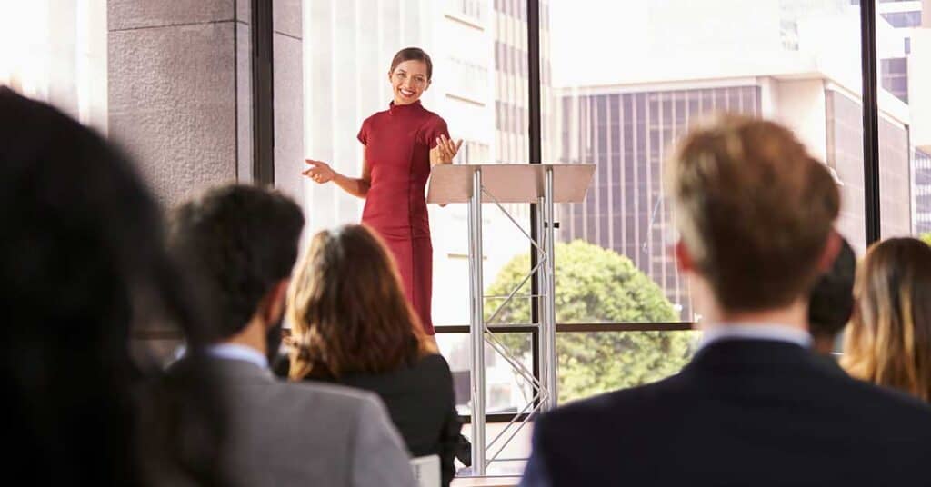 Woman in a red dress stands at a podium, speaking to an audience seated in a modern, sunlit conference room with large windows and others joining online.