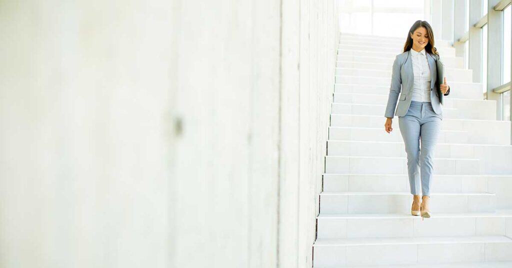 A confident woman in a light gray suit walks down a wide, white staircase in a bright, modern building, exemplifying the strength and elegance of women in contemporary settings.