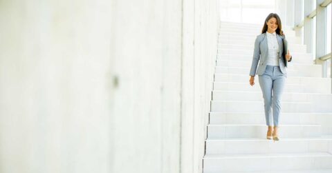 A confident woman in a light gray suit walks down a wide, white staircase in a bright, modern building, exemplifying the strength and elegance of women in contemporary settings.