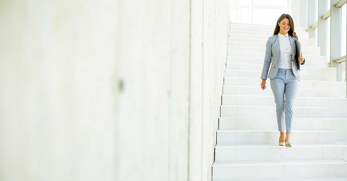 A confident woman in a light gray suit walks down a wide, white staircase in a bright, modern building, exemplifying the strength and elegance of women in contemporary settings.