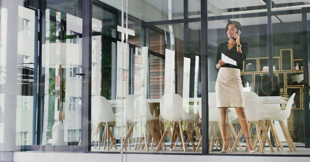 A woman stands in a modern glass-walled office, holding papers and talking on her mobile phone, maintaining her reputation as a dedicated professional; a conference table and chairs are visible in the background.