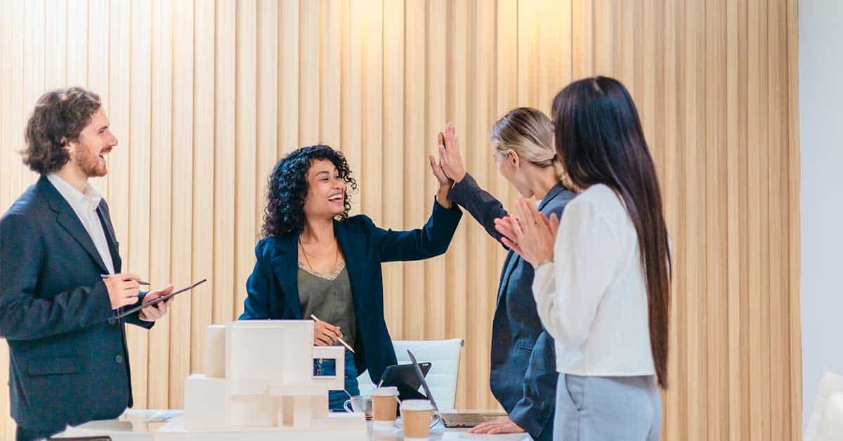Four colleagues stand around a desk in an office. Demonstrating leadership, two women in the center give each other a high five while the others observe and smile.