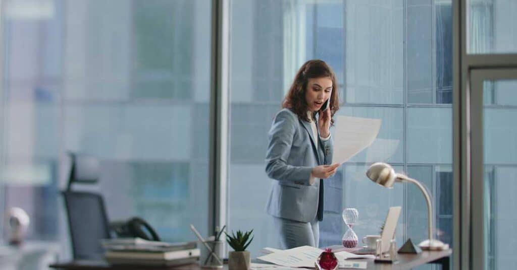 A woman in a business suit stands at a desk in a modern office, talking on the phone and looking surprised while holding a coaching document.