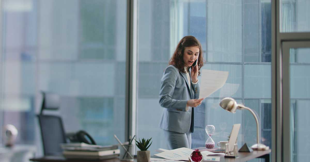 A woman in a business suit stands at a desk in a modern office, talking on the phone and looking surprised while holding a coaching document.