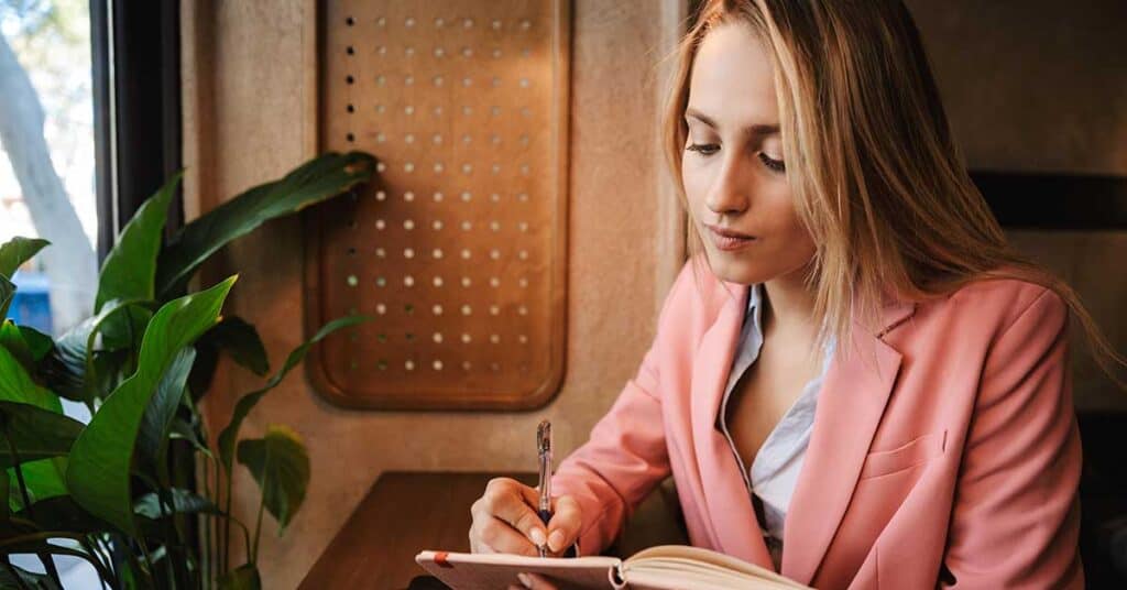 A woman in a pink blazer sits at a table by a window, writing in her notebook as she enjoys a quiet moment of self-reflection beside a green potted plant.
