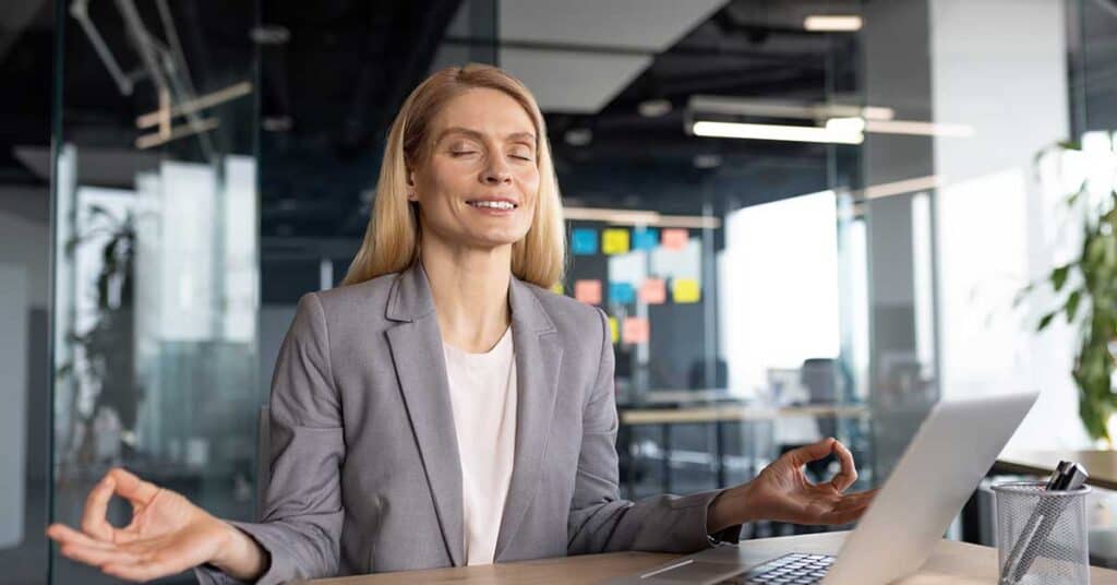 A woman in a gray blazer sits at a desk with a laptop, eyes closed and hands in a meditation pose, embracing work-life balance in a calm, modern office setting.