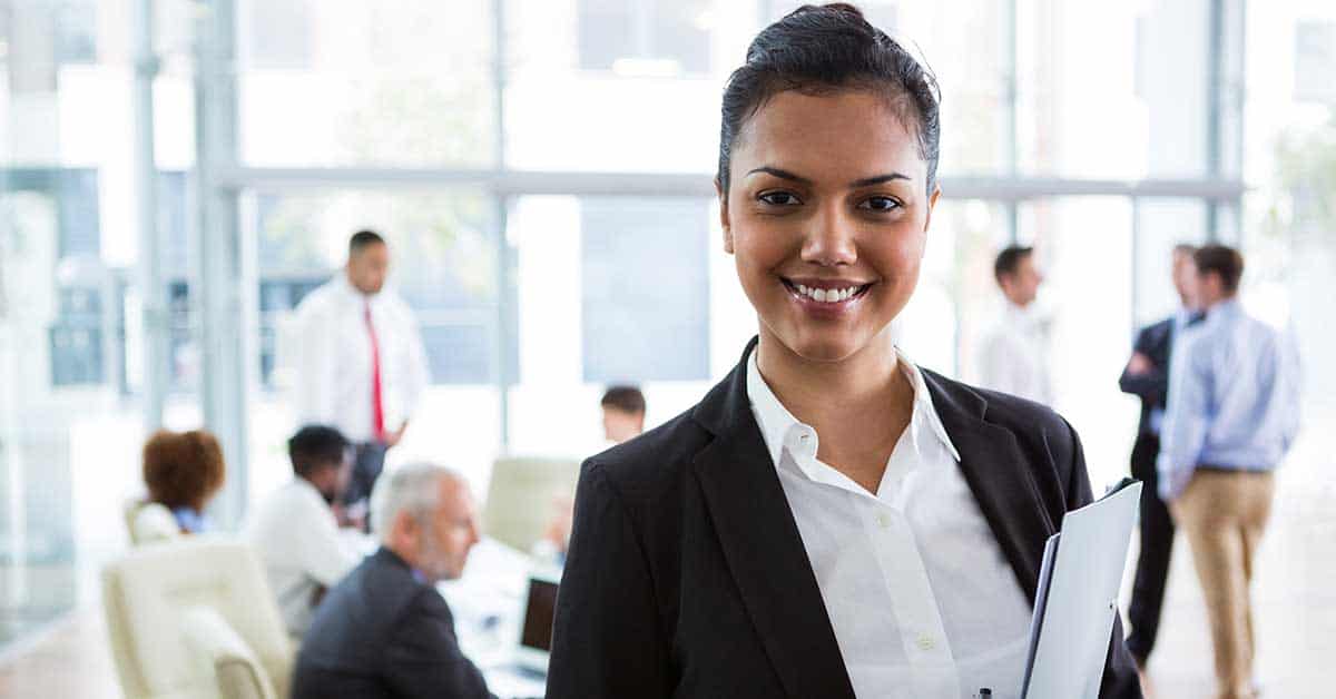 A woman in business attire smiles at the camera while holding documents in an office, ensuring there’s no misalignment as her colleagues work in the background.