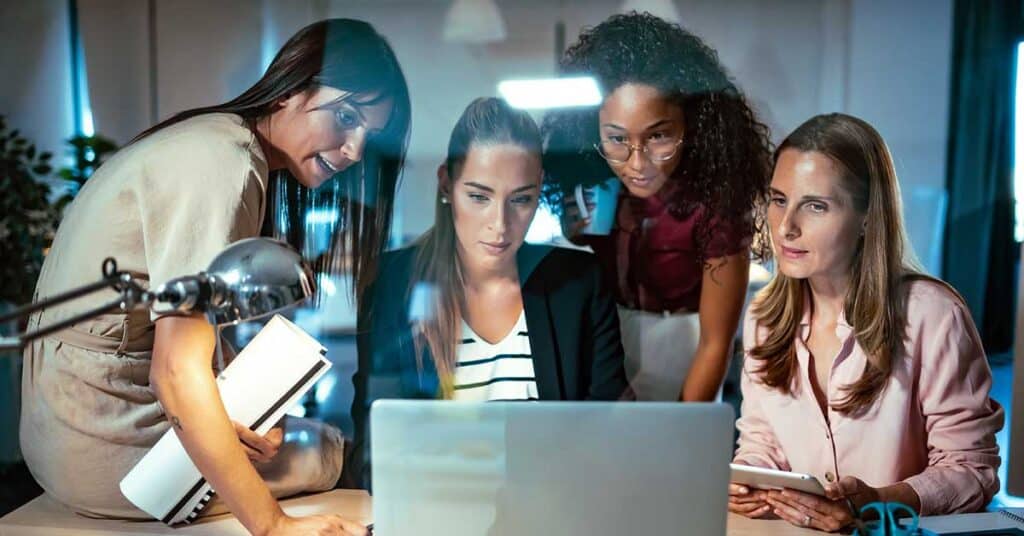 Four women in business attire demonstrate leadership as they collaborate around a laptop at a modern office desk, with documents and a tablet visible.