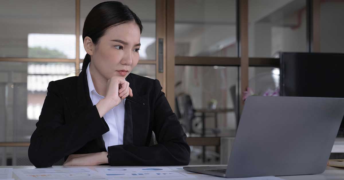 A woman in business attire sits at a desk, looking thoughtfully at a laptop screen displaying AI insights, with documents spread out in front of her.
