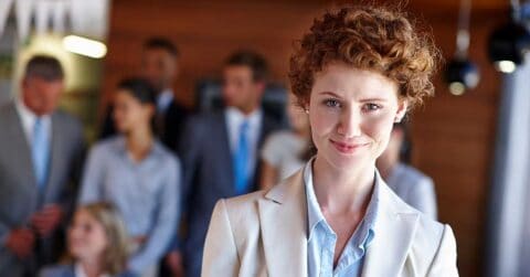 A confident leader in business attire stands in the foreground, smiling, with a group of professionally dressed people blurred in the background.