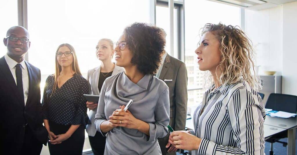 Six professionally dressed people stand in an office, demonstrating leadership as they look attentively in one direction, with windows and city buildings visible in the background.