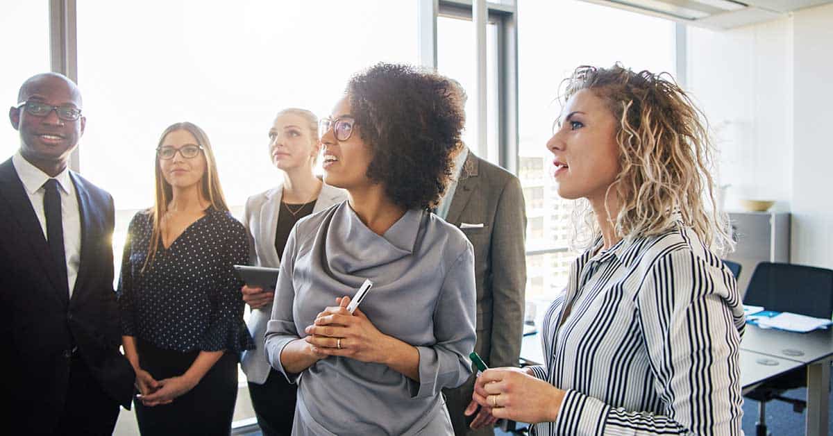 Six professionally dressed people stand in an office, demonstrating leadership as they look attentively in one direction, with windows and city buildings visible in the background.