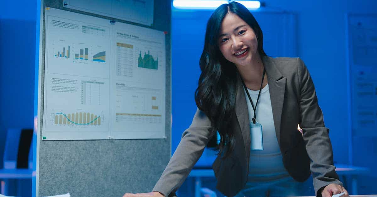 A woman in business attire stands by a board displaying AI-powered charts and graphs, smiling at the camera in an office setting.