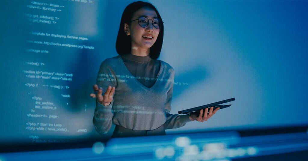 A woman demonstrating leadership holds a tablet as she stands before a projected screen of computer code, appearing to give a presentation.